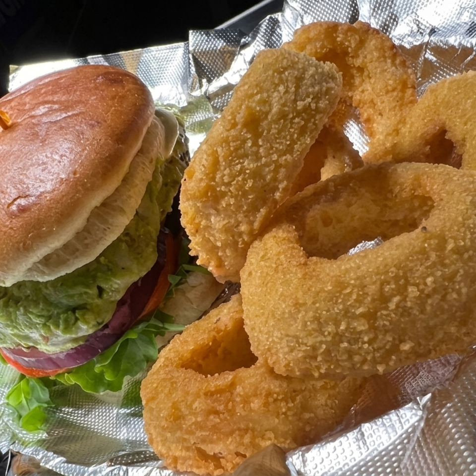 Burger with lettuce, tomato, and onion served alongside crispy breaded onion rings on foil-lined paper.